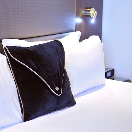 Stamford Plaza Melbourne Hotel Eksteriør billede The photo shows a neatly arranged bed with white linens. There are several pillows, with one black pillow featuring a distinctive design on top of the white pillows. The headboard of the bed is visible, along with a stylish wall-mounted light fixture