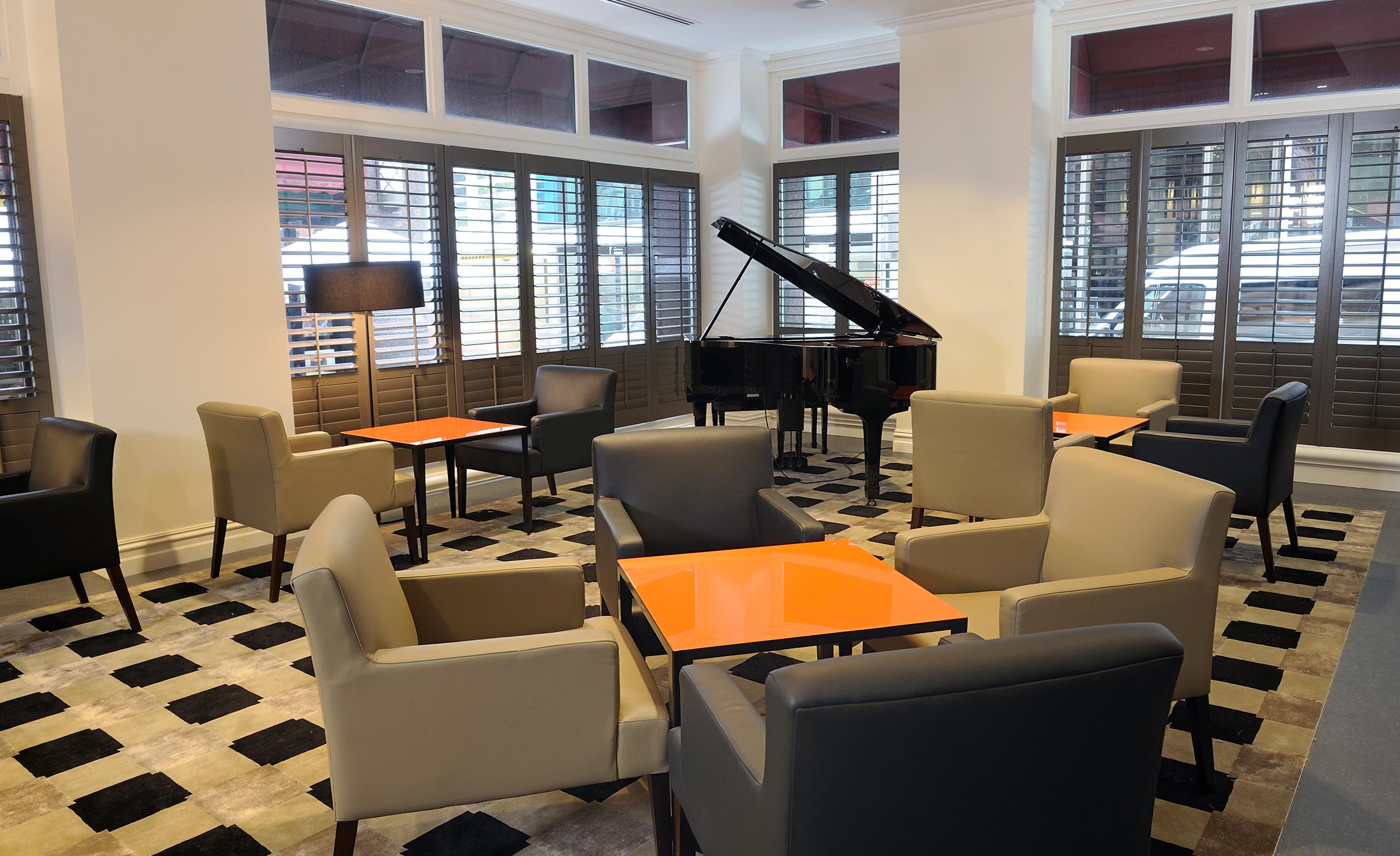 Stamford Plaza Melbourne Hotel Eksteriør billede The photo shows a stylish indoor lounge area. It features several light gray armchairs arranged around orange square tables. In the corner, there is a black grand piano, adding an elegant touch to the space. The room has large windows with wooden shu