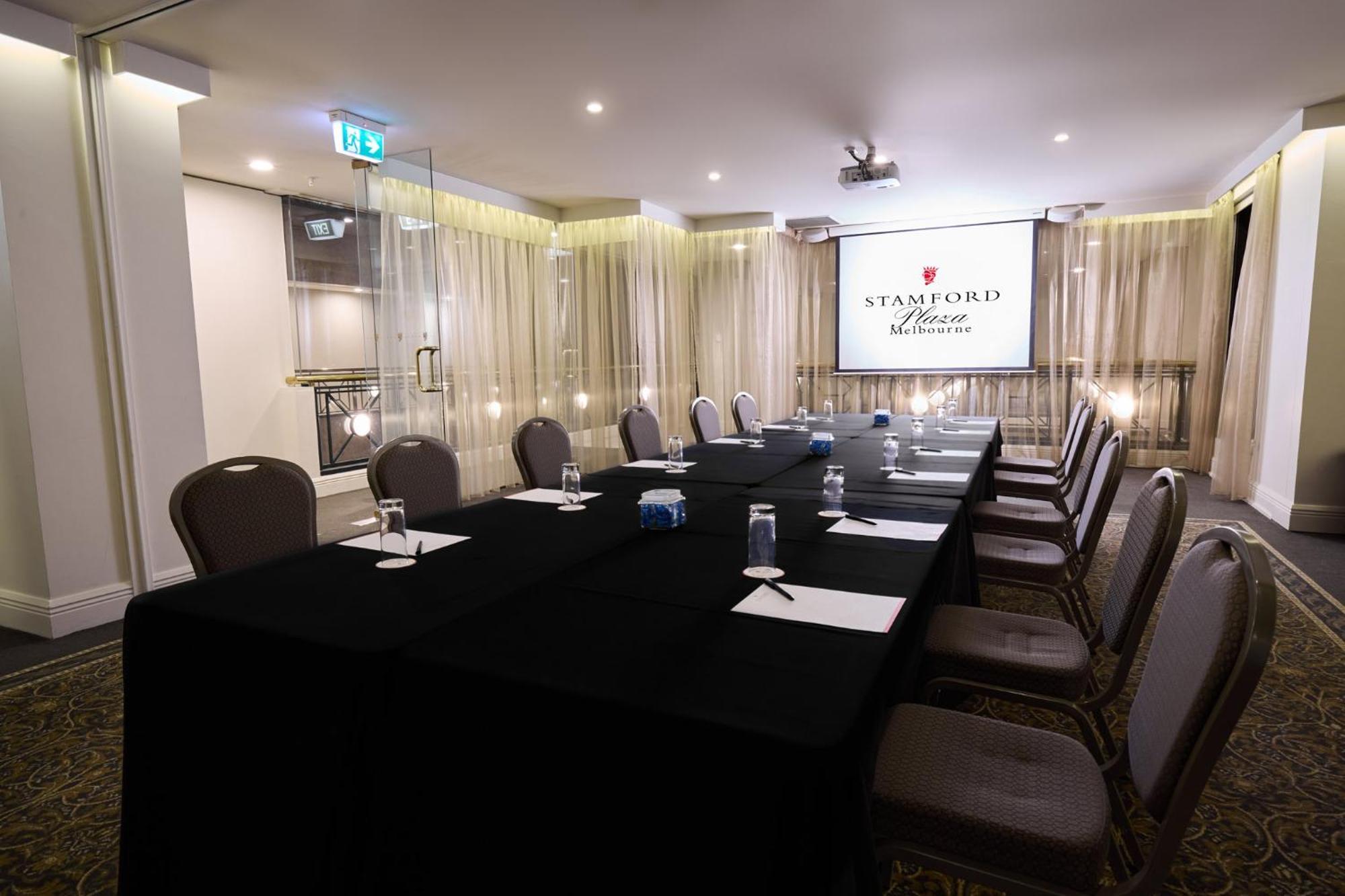 Stamford Plaza Melbourne Hotel Eksteriør billede The photo shows a well-appointed conference room designed for meetings or presentations. There is a long rectangular table covered with a black tablecloth, surrounded by upholstered chairs. On the table, there are several folded pieces of paper and g