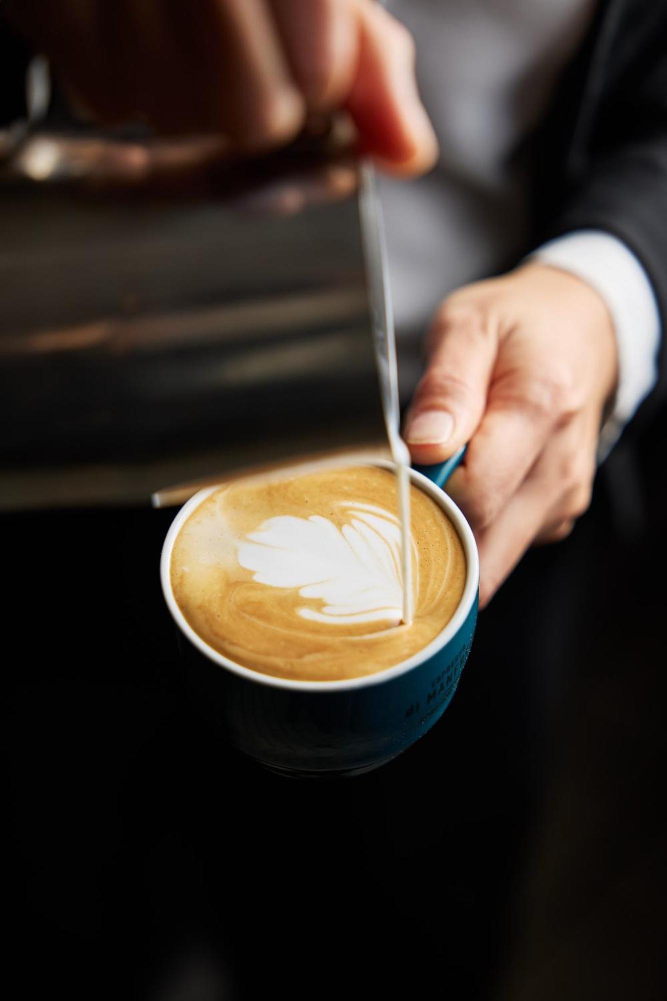 Stamford Plaza Melbourne Hotel Eksteriør billede The photo shows a close-up of a hand holding a cup of coffee with a latte art design on the surface. The coffee appears to be a creamy, caramel color, likely a latte or cappuccino. A small stream of milk is being poured into the cup, creating a heart