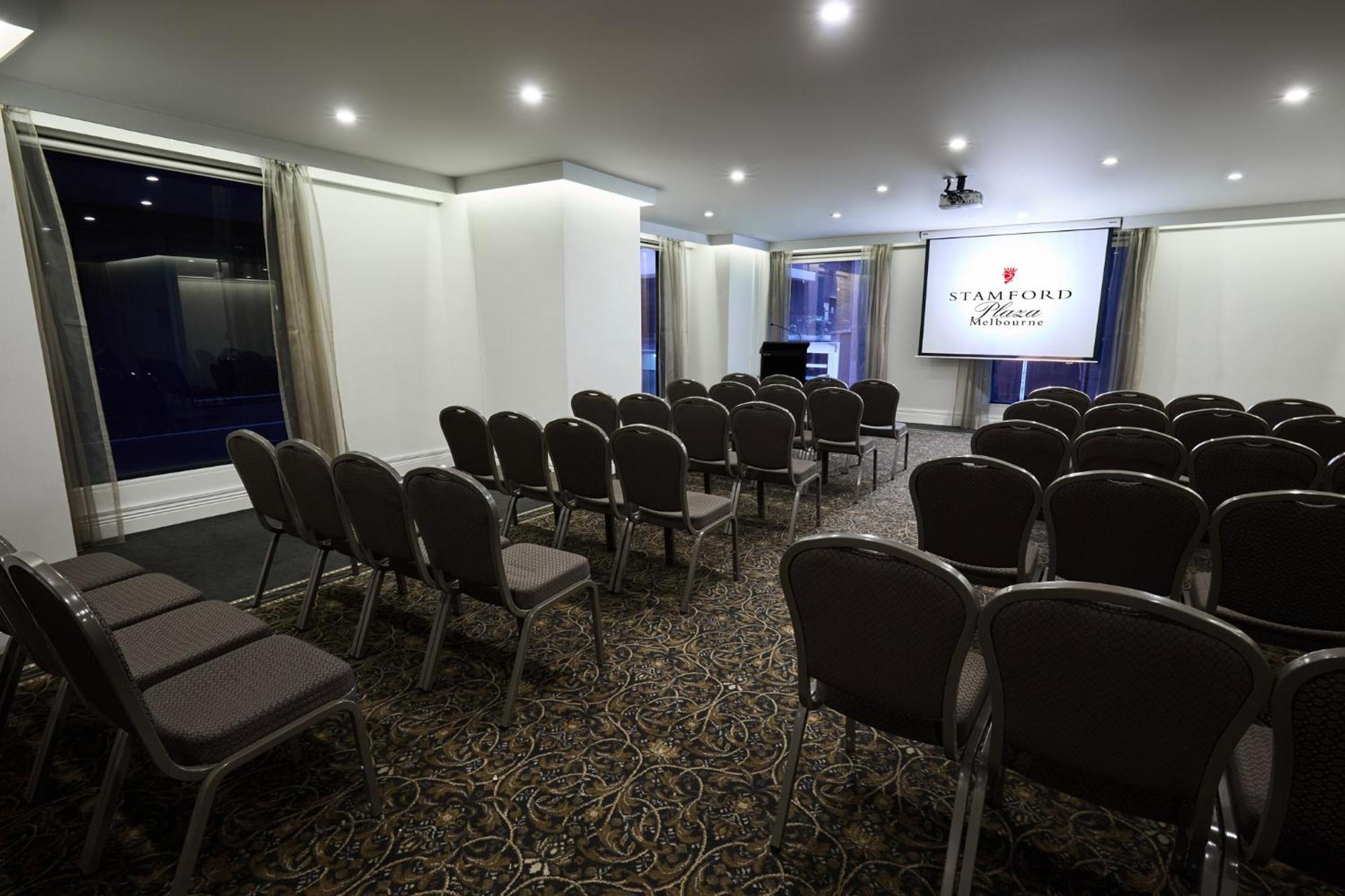 Stamford Plaza Melbourne Hotel Eksteriør billede The image shows a conference or meeting room set up for an event. There are rows of gray chairs arranged facing a large screen displaying the name "Warford House." The room is well-lit with overhead lighting and has large windows covered with light-c