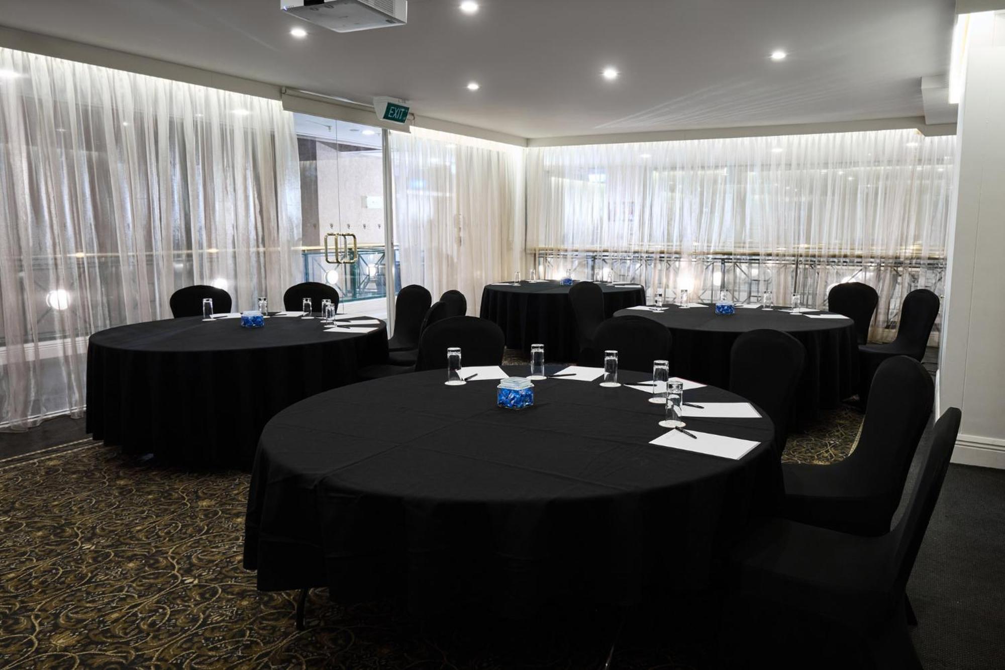Stamford Plaza Melbourne Hotel Eksteriør billede The photo shows a meeting or conference room set up with several round tables covered in black tablecloths. Each table is accompanied by black chairs. There are some white notepads and pens placed on the tables, and a small decorative item, possibly 